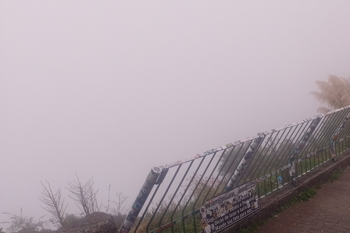 Neblina fechada encobrindo o vale visto de um mirante na serra, com o guarda-corpo em primeiro plano
