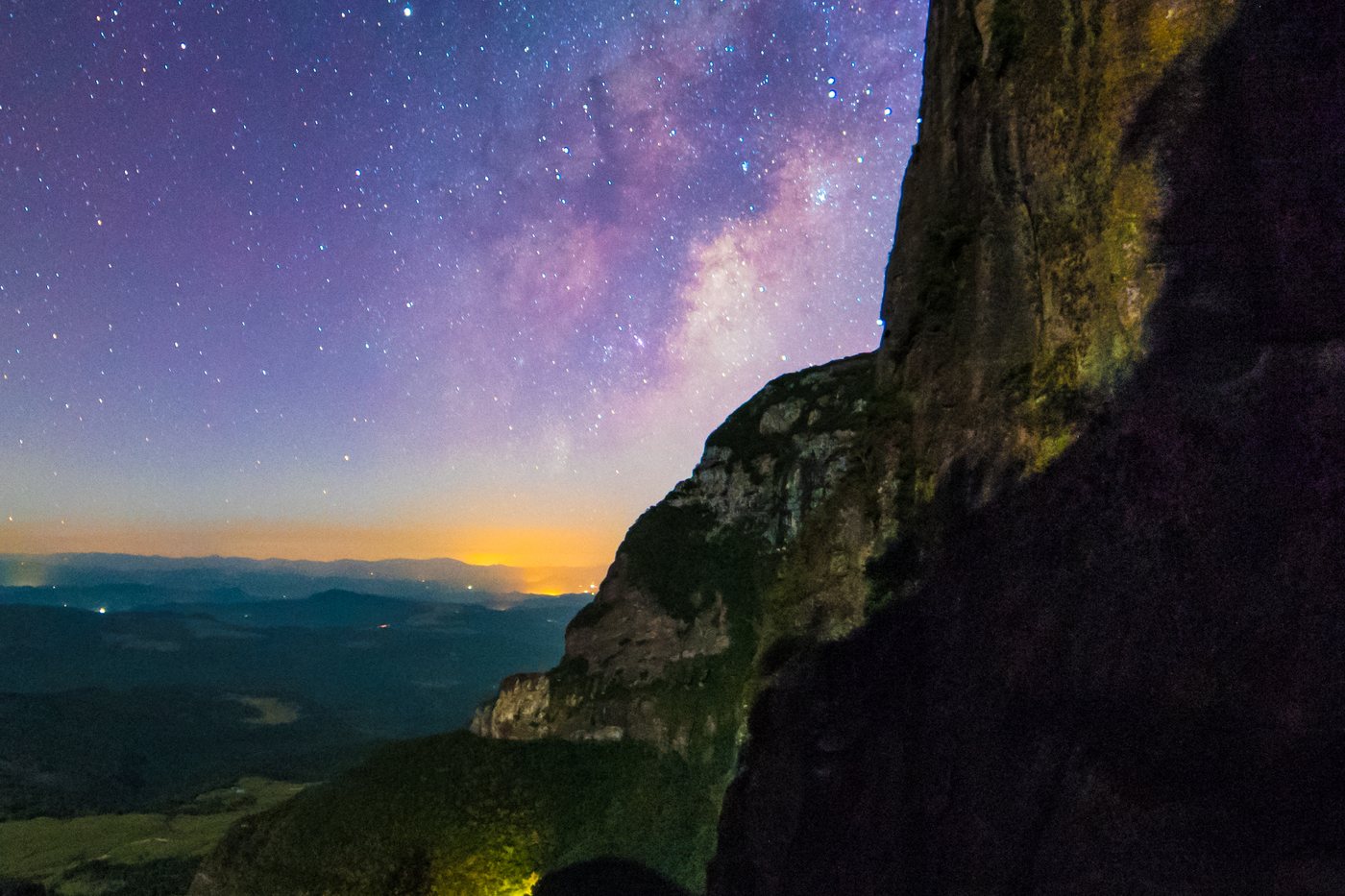 Céu estrelado com a faixa da Via Láctea sobre o paredão de uma serra, com o brilho de um vale iluminado ao fundo